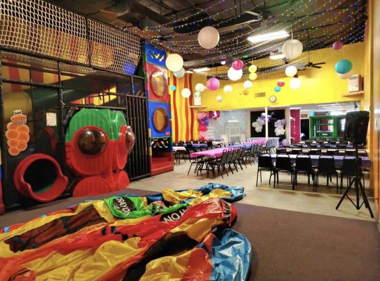 Indoor play structure with tunnels and portholes next to rows of party tables under twinkling string lights